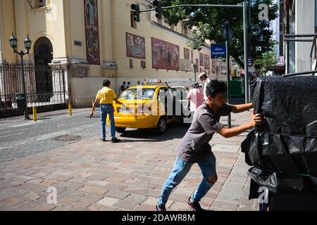 Cordoba, Argentina - January, 2020: Young man hard working pushing heavy cart on the street. Taxi driver dropping of passengers from yellow taxi car. Stock Photo