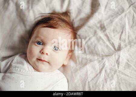 Closeup portrait of cute Caucasian red-haired newborn baby. Adorable funny child infant with blue grey eyes lying on bed looking at camera. Authentic Stock Photo