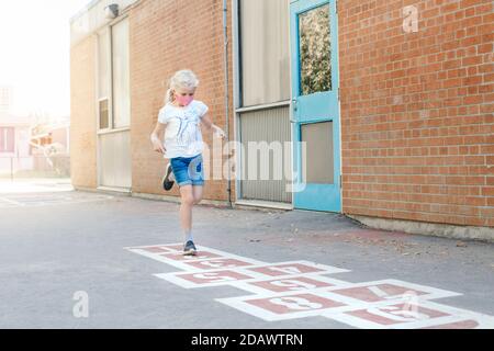 Child girl in face mask playing jumping hopscotch on school yard. Funny activity game for kids on playground outdoor. Street sport for children. Stock Photo