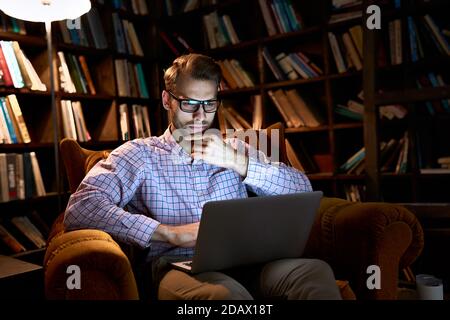 Serious young business man using laptop working late online sitting in chair. Stock Photo