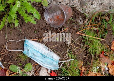 A light blue discarded medical face mask lying on dirt. Grass and plant present, and empty plastic drink cup above mask. Room for text. Stock Photo