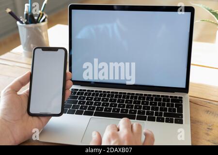 Man using computer holding smartphone with white empty mockup screen Stock Photo