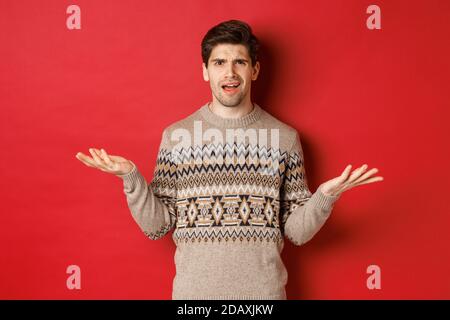 Portrait of confused and disappointed handsome guy, complaining about christmas, spread hands sideways and frowning displeased, standing in xmas Stock Photo