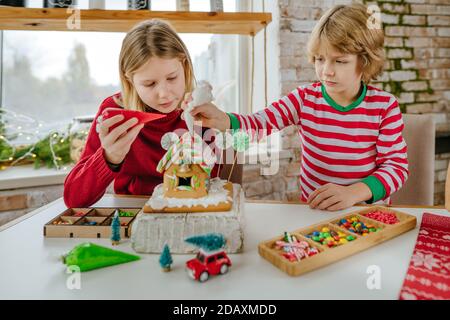 Children decorated Christmas gingerbread house with colorful frosting and candies on the table in the kitchen. Selective focus. Stock Photo
