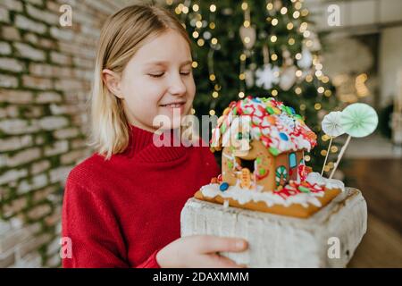 Girl holding Christmas gingerbread house decorated with candies and icing. Christmas family home activities concept. Stock Photo