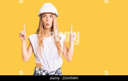 Beautiful caucasian woman with blonde hair wearing hardhat and painter clothes amazed and surprised looking up and pointing with fingers and raised ar Stock Photo