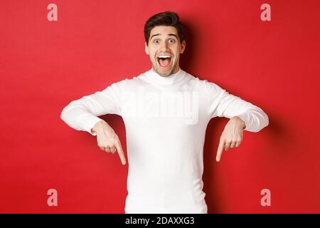 Image of amazed handsome guy in white sweater, reacting excited to christmas advertisement, pointing fingers down, showing logo, standing over red Stock Photo