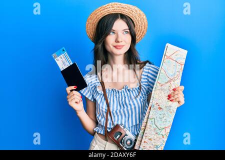 Young beautiful caucasian girl holding passport and city map smiling looking to the side and staring away thinking. Stock Photo