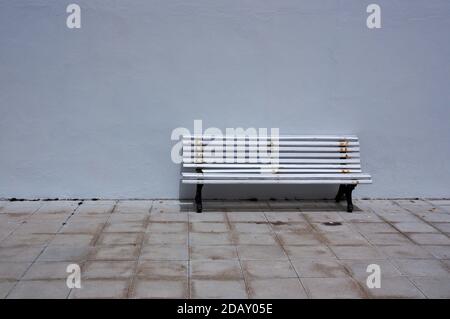 Image of a bench on the street that remains empty next to a white wall on a sidewalk without traffic Stock Photo