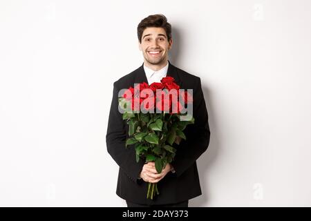 Image of handsome man in black suit, holding bouquet of roses and smiling, standing against white background Stock Photo