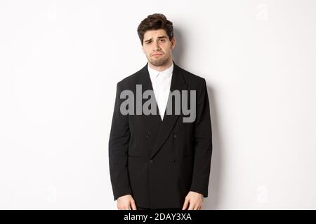 Image of gloomy and sad handsome male model in black suit, frowning and looking tired at camera, standing against white background Stock Photo