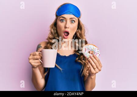 Young blonde girl wearing sleep mask and pyjama having breakfast afraid and shocked with surprise and amazed expression, fear and excited face. Stock Photo