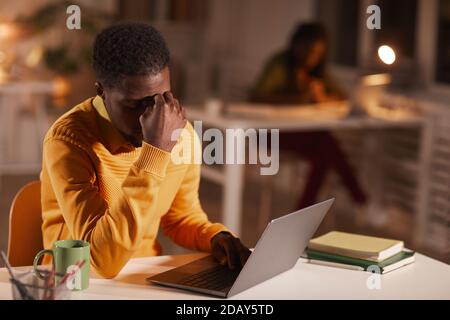 Portrait of exhausted African-American man rubbing eyes while working late at night in dark office, copy space Stock Photo