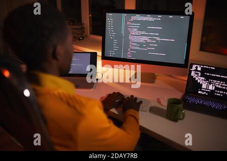 Back view portrait of African-American man writing code while working with multiple computer screens in dark room Stock Photo