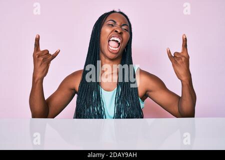 African american woman with braids wearing casual clothes sitting on the table shouting with crazy expression doing rock symbol with hands up. music s Stock Photo