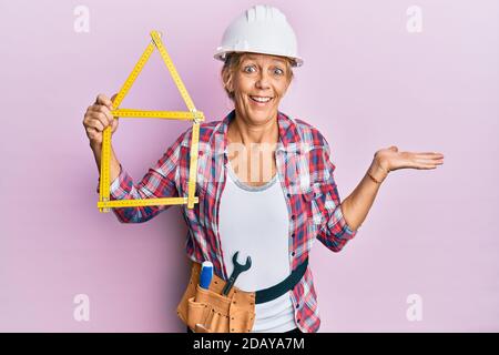 Middle age blonde woman wearing architect hardhat holding build project celebrating achievement with happy smile and winner expression with raised han Stock Photo