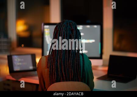Back view portrait of female IT developer writing code on multiple computer screens while working late in dark office, copy space Stock Photo