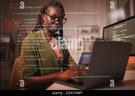 Portrait of contemporary African-American woman using laptop with computer code overlay, copy space Stock Photo