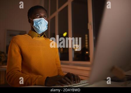 Low angle portrait of young African-American man wearing mask in office while using computer lit by screen in dark, copy space Stock Photo