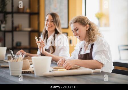 Female potters during the working process in the clay workshop. Woman masters prepare ceramic and clay products at big wooden table Stock Photo