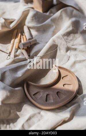 A close-up view of clay plates and a paint brush on a wooden table and dried flowers on a background. Crafted tableware in a pottery studio. Handmade Stock Photo