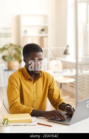 Vertical portrait of contemporary African-American man using laptop while sitting at white desk in office, copy space above Stock Photo