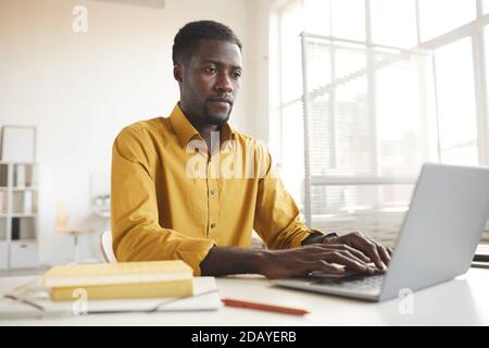 Low angle view at contemporary African-American man using laptop while working at desk in minimal office interior, copy space Stock Photo