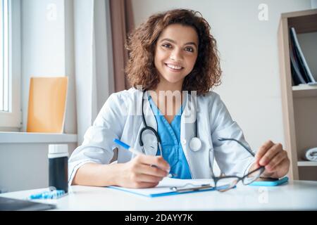 Portrait of female doctor sitting on work desk and smiling at camera. Stock Photo