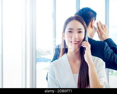 Business Asian couple talking to mobile phone. Young beautiful smiling woman talking with cell phone and standing back with businessman in suit on hug Stock Photo