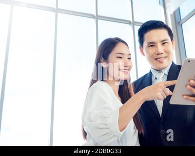 Business couple working together. Asian businessman in suit and smiling young woman pointing at digital tablet while both watching at screen on glass Stock Photo