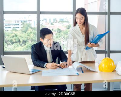 Asian male chief architect or engineer in suit and young female secretary discuss on blueprint with laptop computer and white and yellow hard hats on Stock Photo