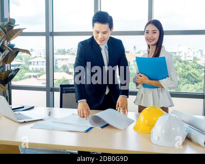 Asian male chief architect or engineer in suit and young female secretary discuss on blueprint with laptop computer and white and yellow hard hats on Stock Photo