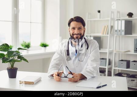 Happy bearded doctor sitting at table in medical office. Stock Photo