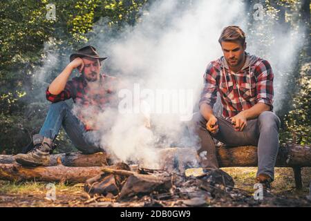 Group of friends enjoying picnic in the forest and drinking beer. Group of two male friends camping with marshmallows over a camp fire. Man doing mans Stock Photo