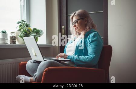 Side view photo of a caucasian senior woman typing something at the laptop while wearing glasses and casual clothes in an armchair Stock Photo