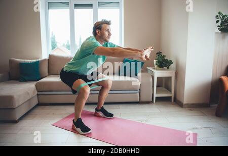 Caucasian man is doing fitness at home using special bands on the floor while squatting Stock Photo