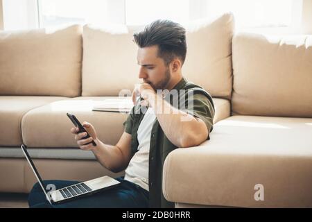 Thoughtful caucasian man is using a phone and laptop in the floor near a sofa while working remotely Stock Photo
