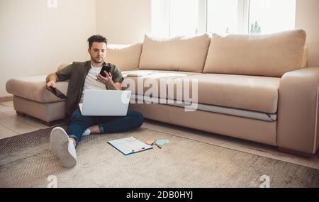 Caucasian bearded man with a phone is lying on floor using a laptop while working remotely online Stock Photo