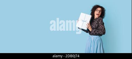 Curly haired woman is showing at camera a present box posing happily near blue free space Stock Photo