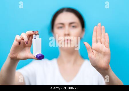 A woman holds an inhaler with medicine in her hand and makes a stop gesture with her hand. Portrait in a blur on a blue background. The concept of ref Stock Photo