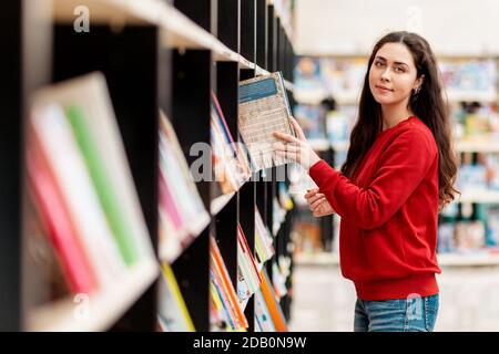 A young Caucasian woman pulls a book from the shelf and smiles sweetly. Blurred shelves of books. The concept of education and purchase of books. Stock Photo