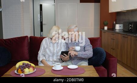 Senior old couple grandparents talking, using digital tablet computer at home. Weekend lifestyle, elderly grandmother and grandfather doing internet shopping choose sale offers together, drinking tea Stock Photo