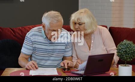 Worried senior retired couple checking calculating bills bank loan payment doing paperwork discuss unpaid debt taxes at home. Stressed old mature grandparents family upset about money problem concept Stock Photo