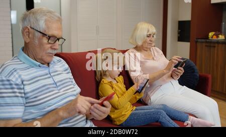 Senior couple grandparents with child girl granddaughter spending time home together, sitting on sofa in living room, using digital tablet, mobile phone. Look concentrated and antisocial Stock Photo
