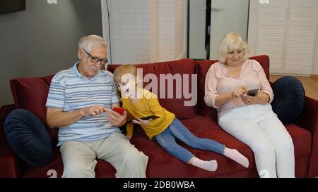 Senior couple grandparents with child girl grandchildren spending time home together, sitting on sofa in living room, using digital tablet, mobile phone. Watching videos, playing games, social network Stock Photo
