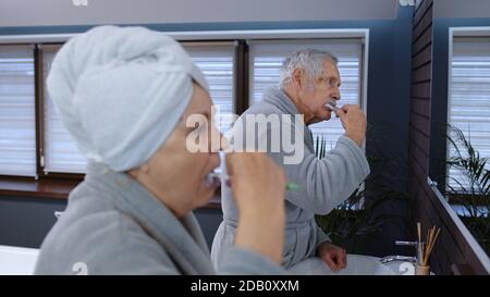 Cheerful old senior couple grandparents man and woman brushing teeth and looking into a mirror. Elderly grandmother and grandfather doing morning hygiene at luxury bathroom at home Stock Photo