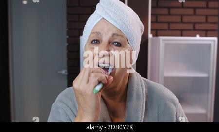 Old senior woman grandmother with towel in bathrobe brushing teeth and looking into a mirror. Elderly grandma doing morning hygiene after shower at luxury bathroom at home. Point of view. POV shot Stock Photo