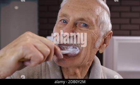 Attractive old senior man grandfather in bathrobe brushing teeth looking into mirror. Handsome elderly grandpa doing morning hygiene after shower at luxury bathroom at home. Point of view. POV shot Stock Photo