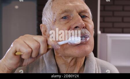 Cheerful old senior man grandfather in bathrobe brushing teeth and looking into mirror. Handsome elderly grandpa doing morning hygiene after shower at luxury bathroom at home. Point of view. POV shot Stock Photo