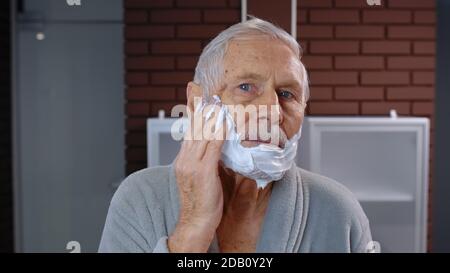 Attractive old senior man grandfather in bathrobe applying foam to face for shaving and looking into mirror. Handsome elderly grandpa doing morning skin hygiene after shower at bathroom home. POV shot Stock Photo